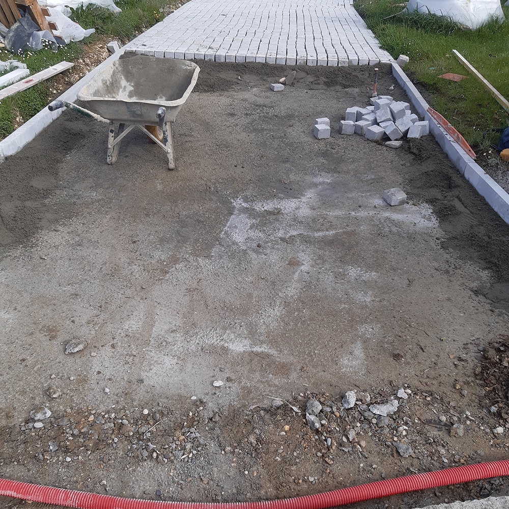 Construction d'Allée en Pavés : Guide et Étapes de Pose Brouette et pavés sur un lit de sable pour la construction d'une allée de jardin bordée de bordures en béton.