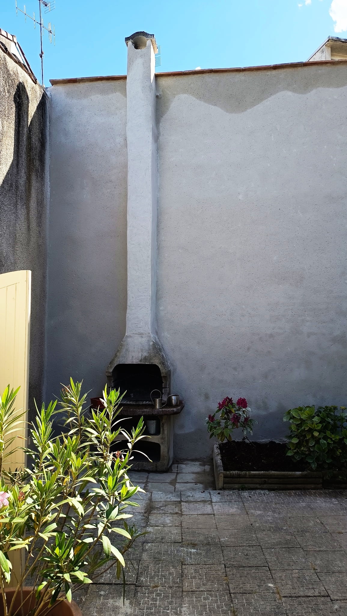 Barbecue en pierre avec une haute cheminée blanche dans un patio ensoleillé entouré de plantes vertes.