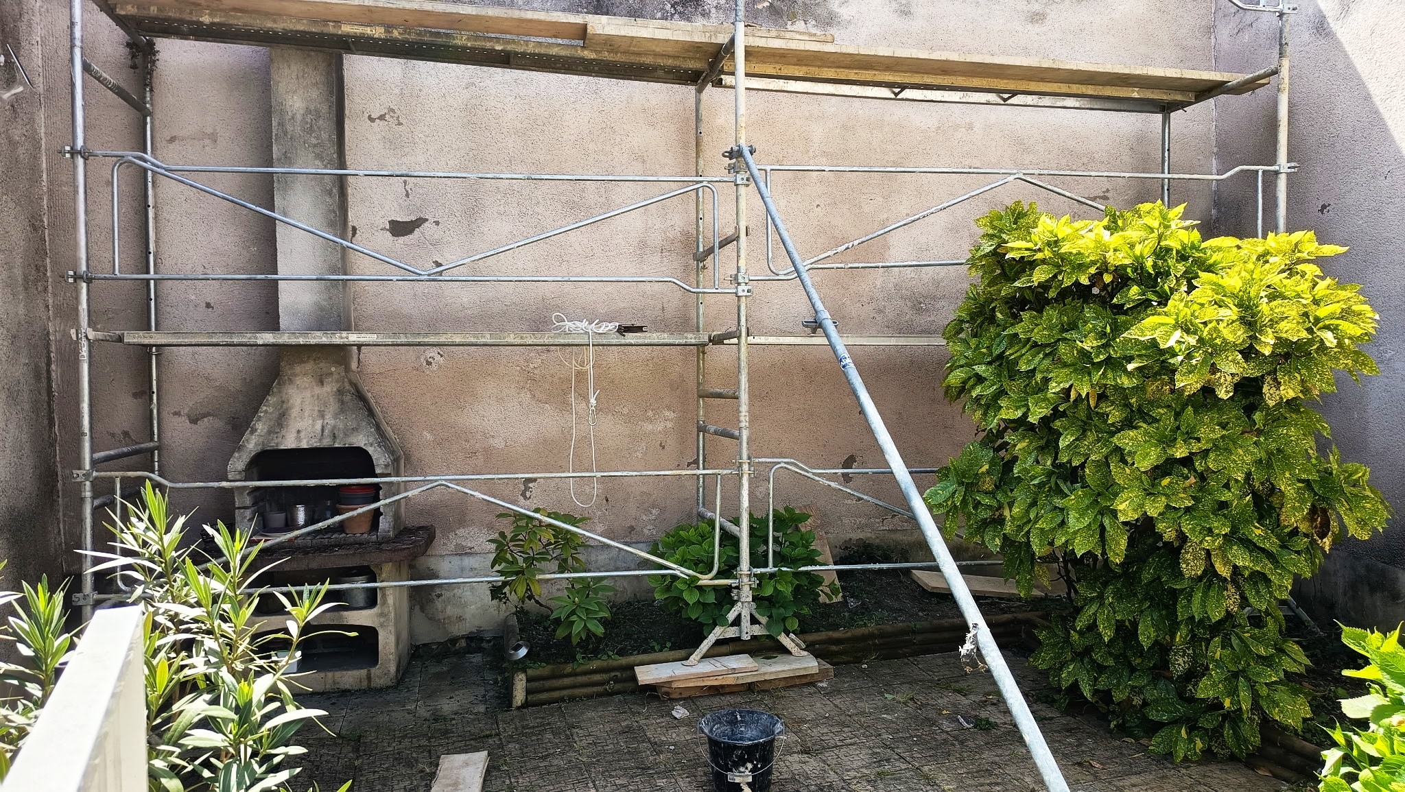 Échafaudage métallique sur une terrasse avec cheminée extérieure, arbustes verts et seau de chantier.