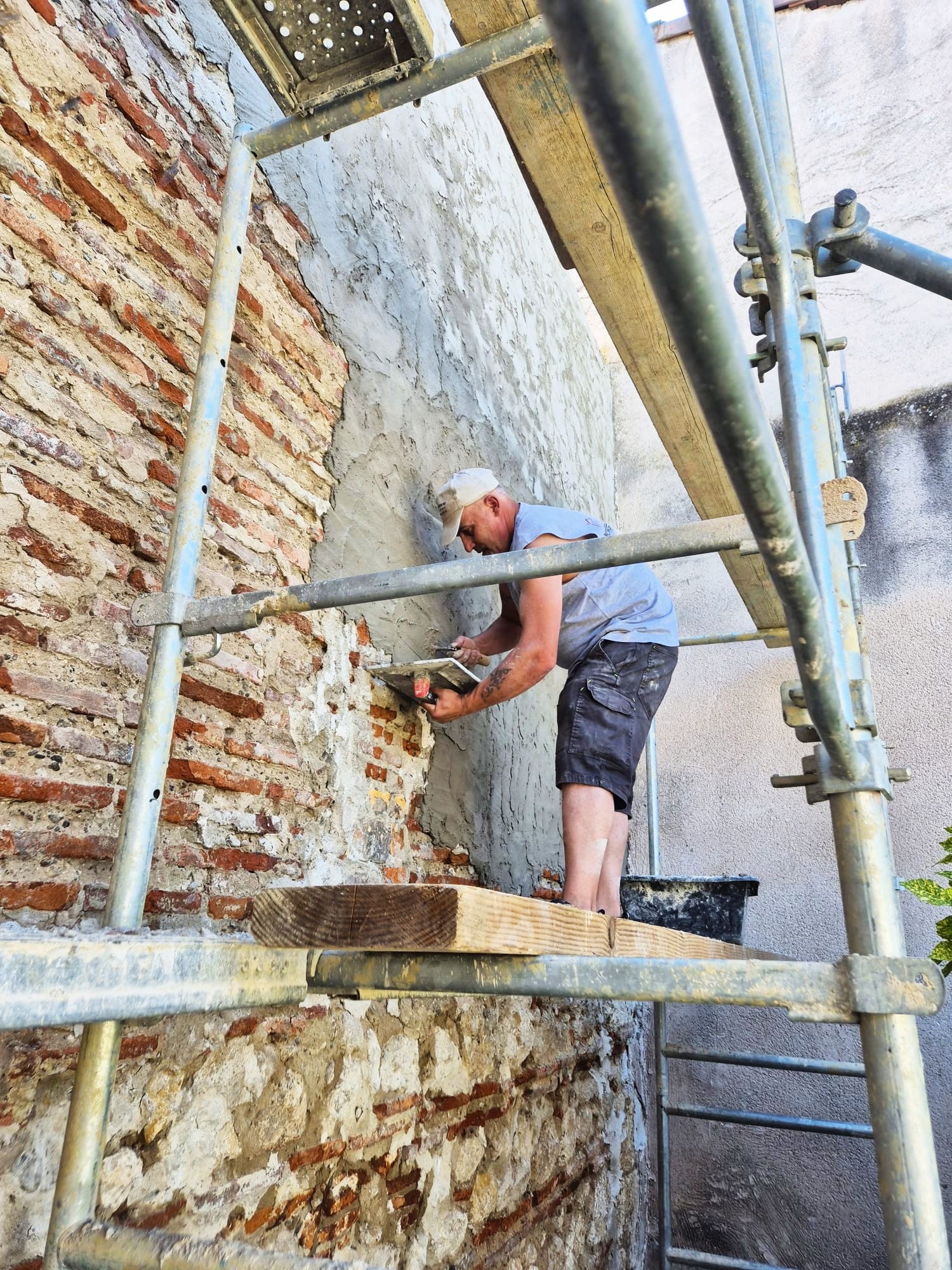 Maçon appliquant du mortier sur un mur en briques anciennes depuis un échafaudage de chantier.