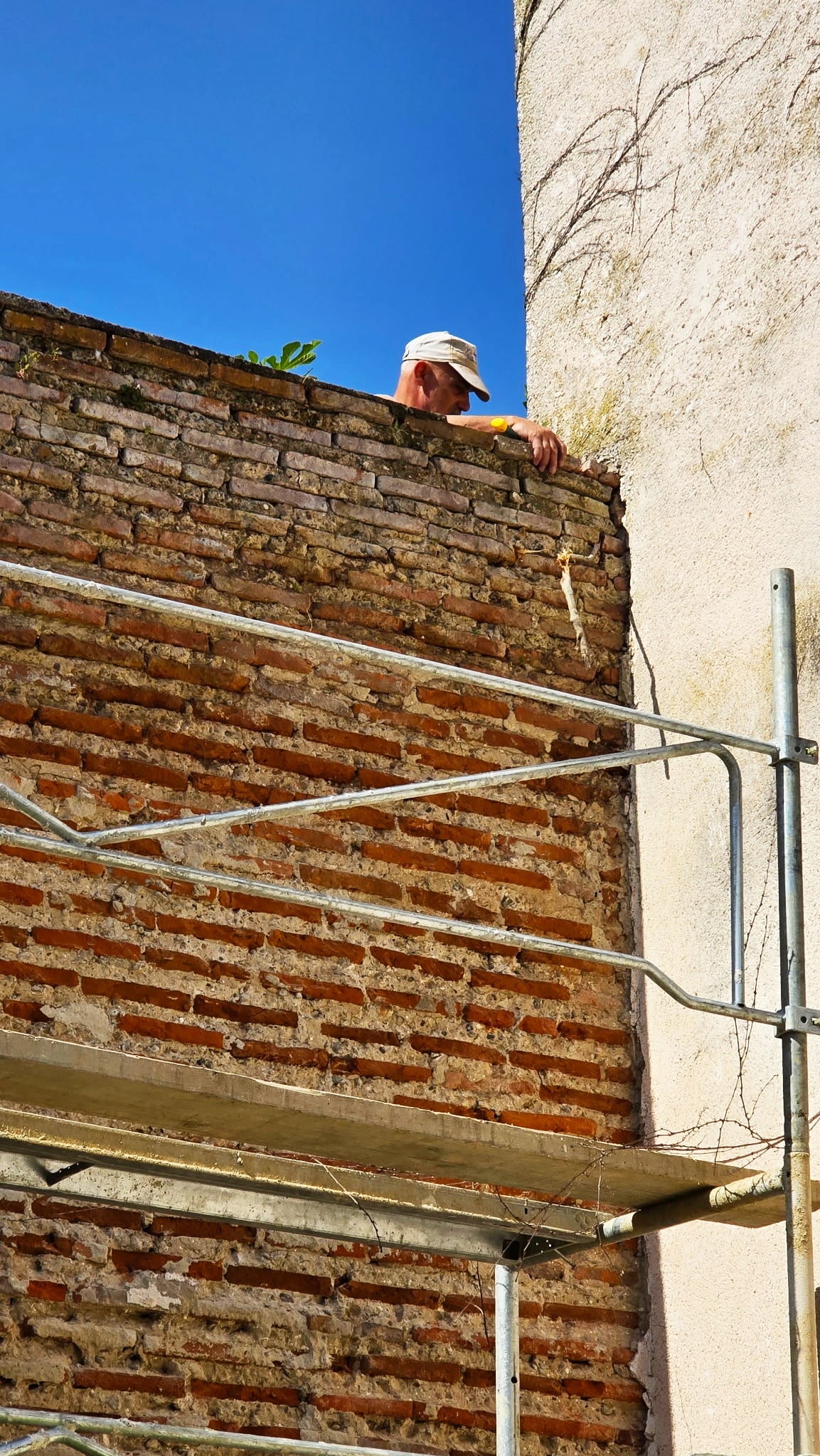 Maçon sur un échafaudage inspectant un mur de briques anciennes en rénovation sous un ciel bleu.